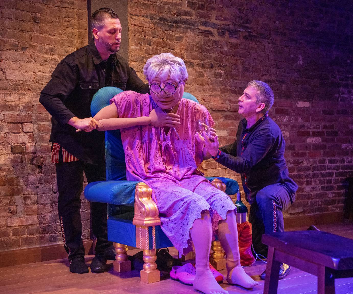 Puppet Sonia, seated on her torquoise padded chair, brings her hand to her chest while she is coughing as a puppet handler and her nephew, Fred try to help.